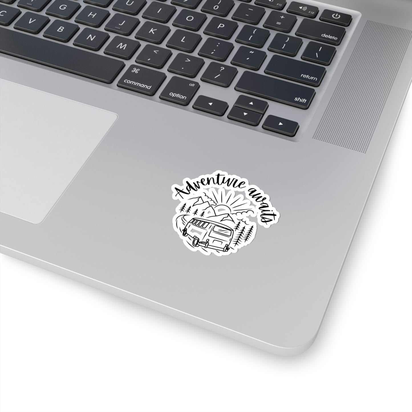 Sticker on a laptop with a drawing of a campervan in the mountains and words "adventure awaits" on white background.