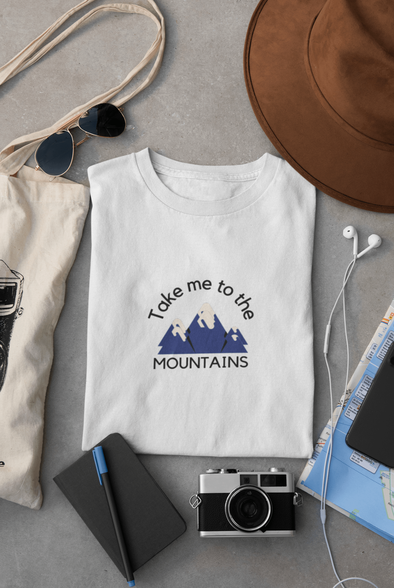 White graphic T-shirt with words "take me to the mountains" printed on the front, surrounded by travel gear.