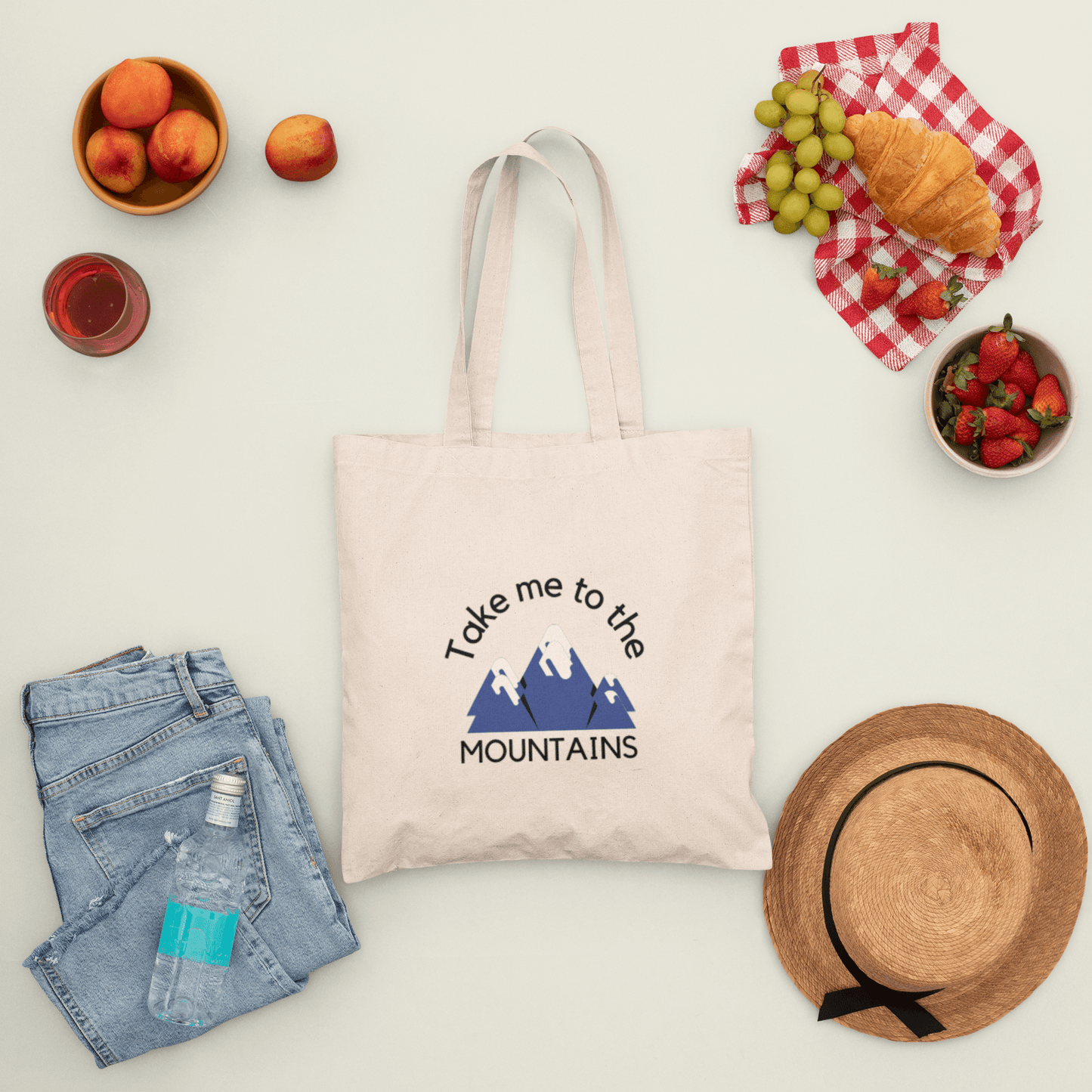 Canvas tote bag with print "take me to the mountains", surrounded by picnic food, a hat, and jeans.