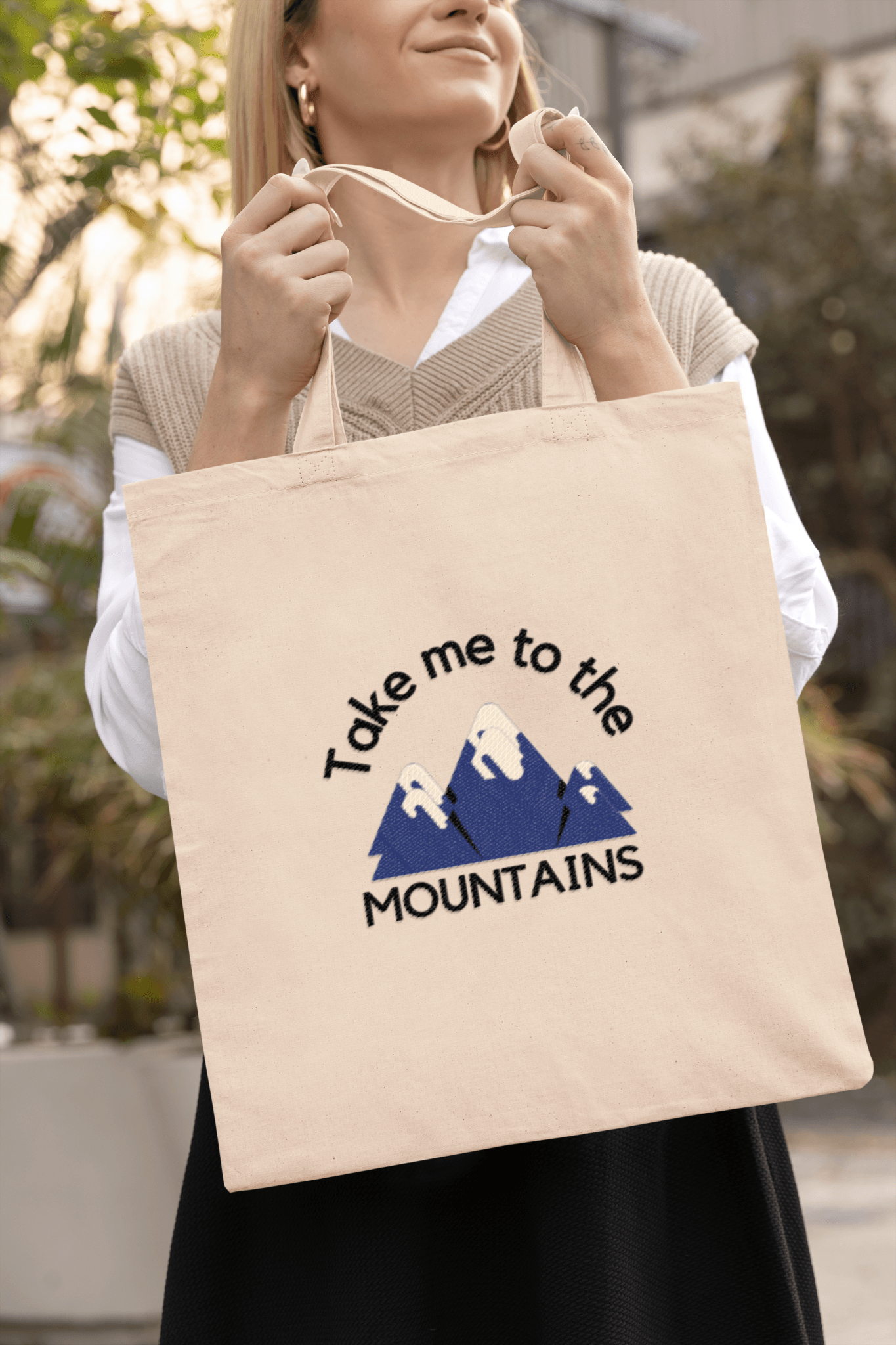 Smiling woman holding a canvas tote bag with print "take me to the mountains".